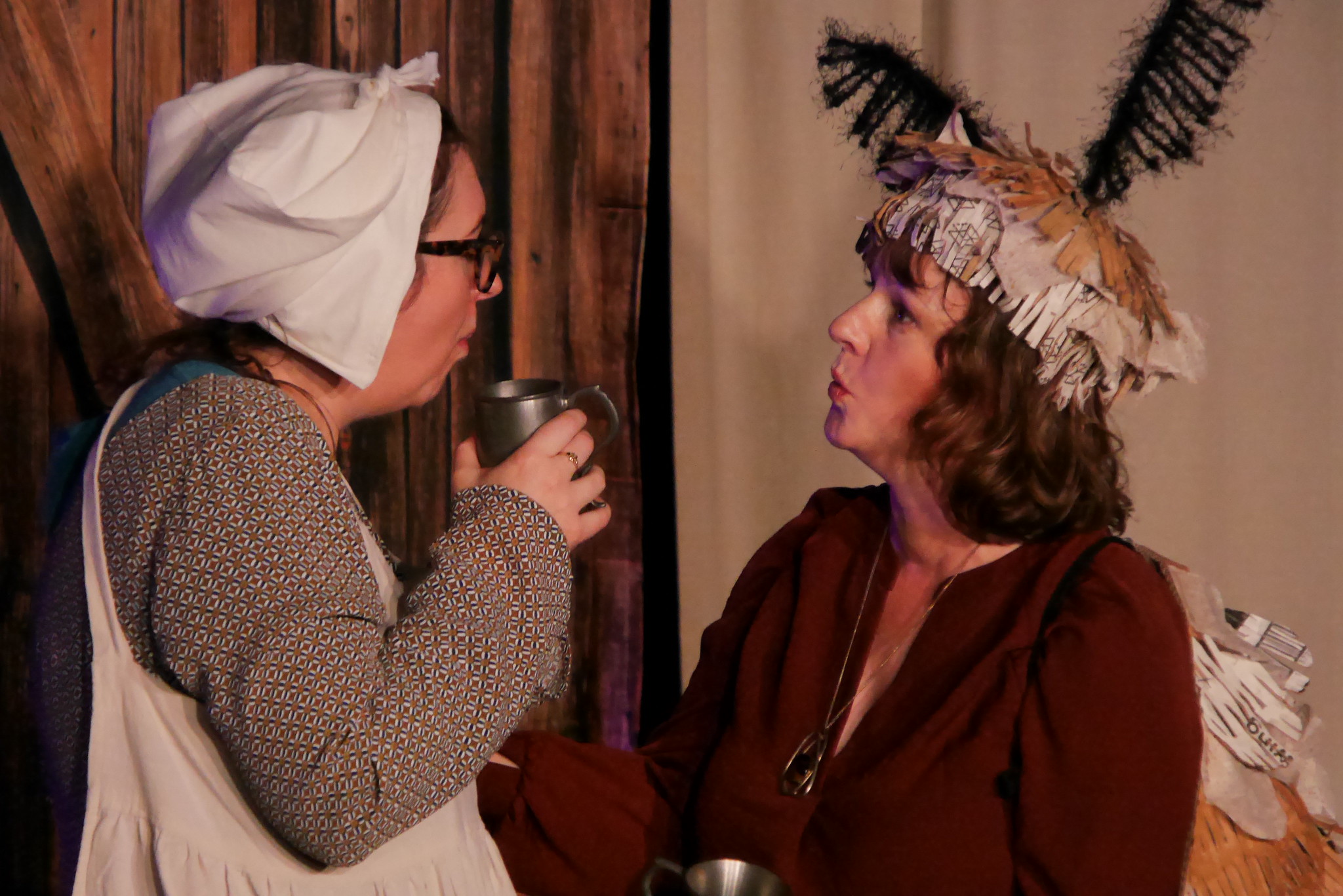 A woman in an apron and bonnet talks over a tankard of ale with her half-moth friend